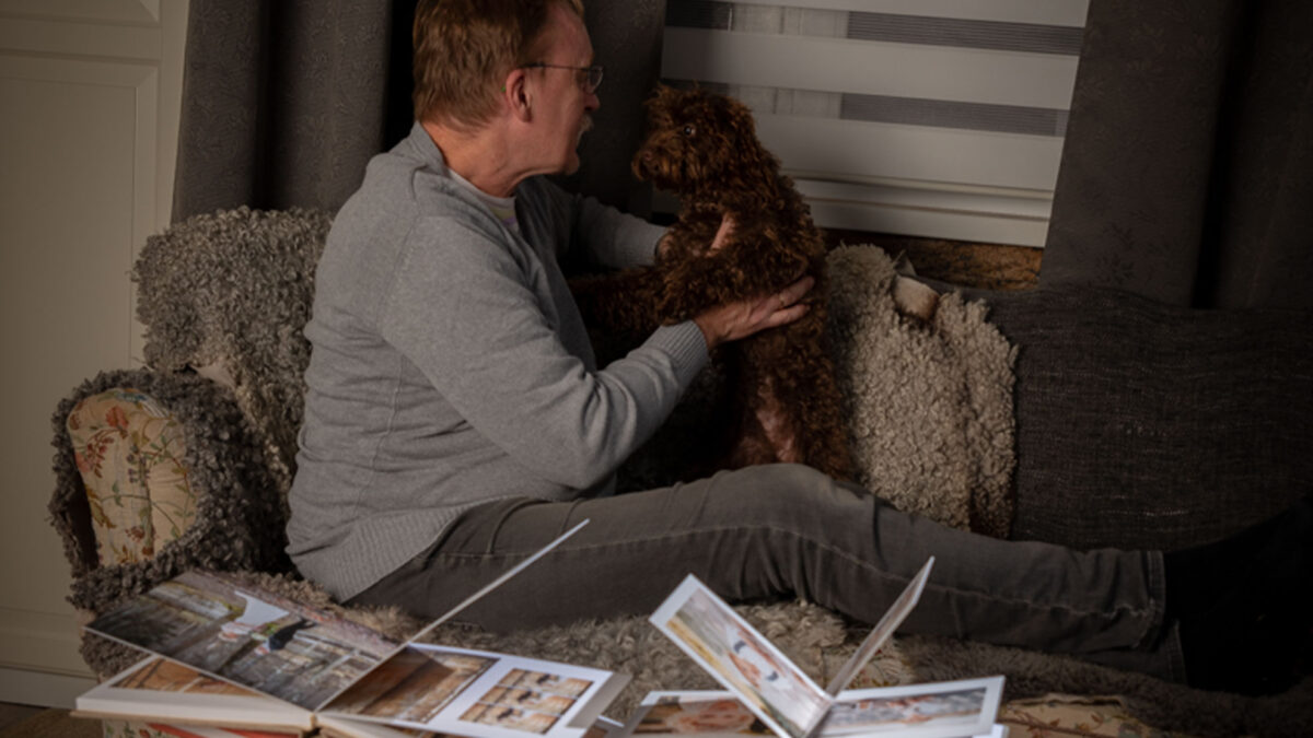En man sitter i en soffa han pratar med en brun hund, på soffbordet ligger fotoböcker