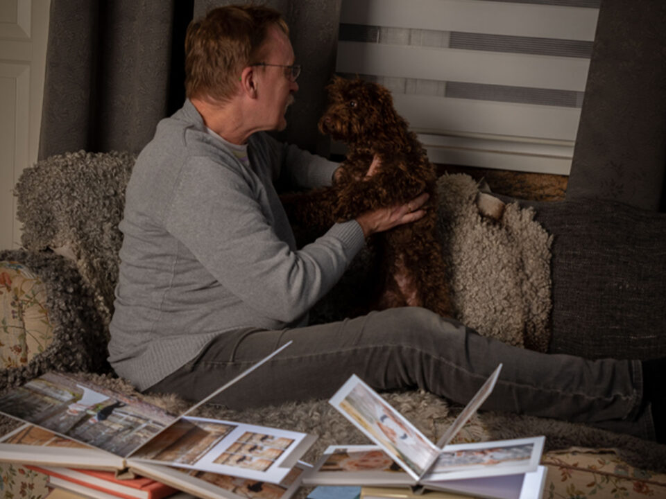 En man sitter i en soffa han pratar med en brun hund, på soffbordet ligger fotoböcker