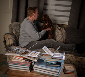En man sitter i en soffa han pratar med en brun hund, på soffbordet ligger fotoböcker